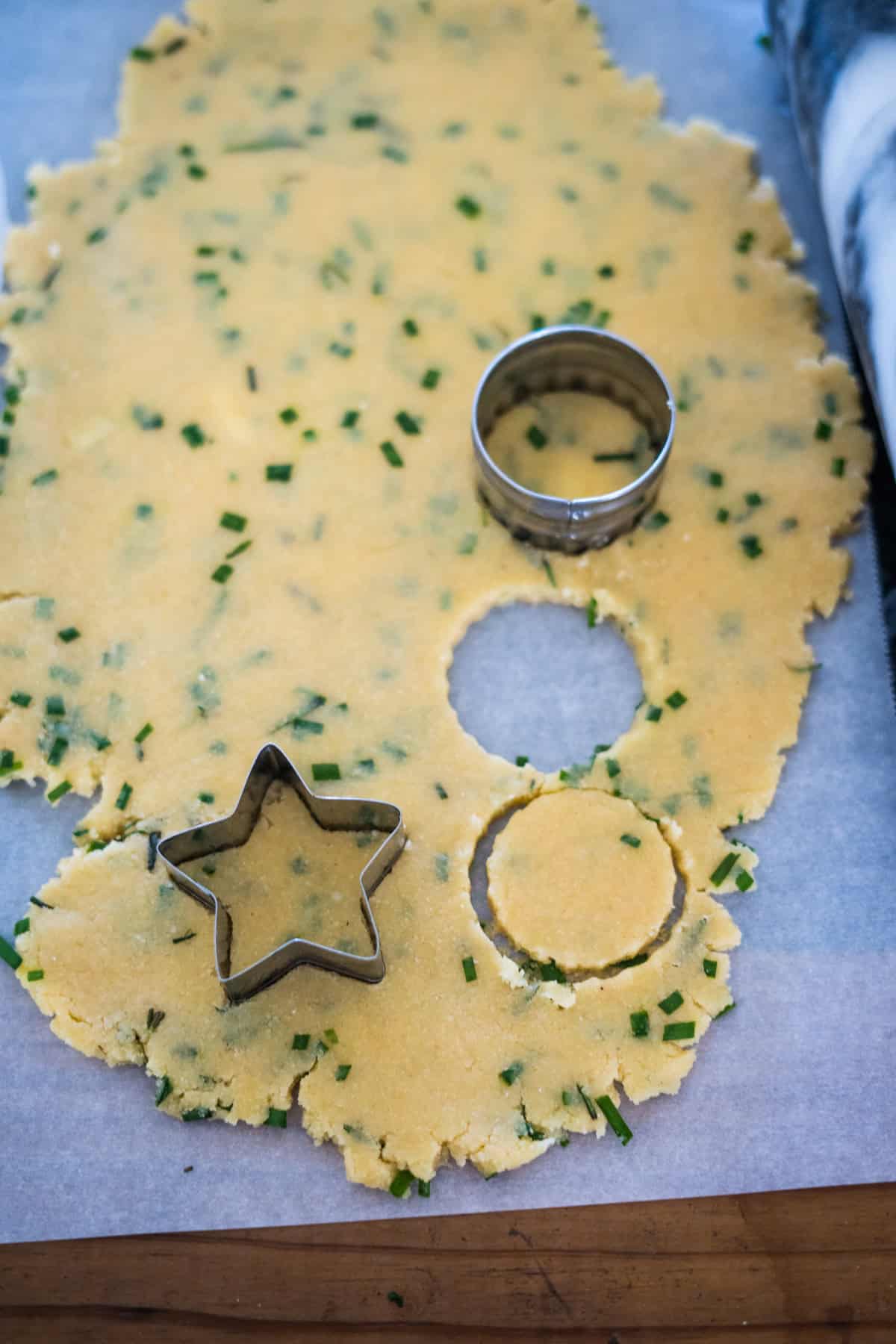 Rolled out dough with chives and hints of cheese, on parchment, featuring cut-out shapes of a circle and a star using metal cookie cutters. A marble rolling pin is partially visible on the side, ready to craft these delightful cheese crackers.