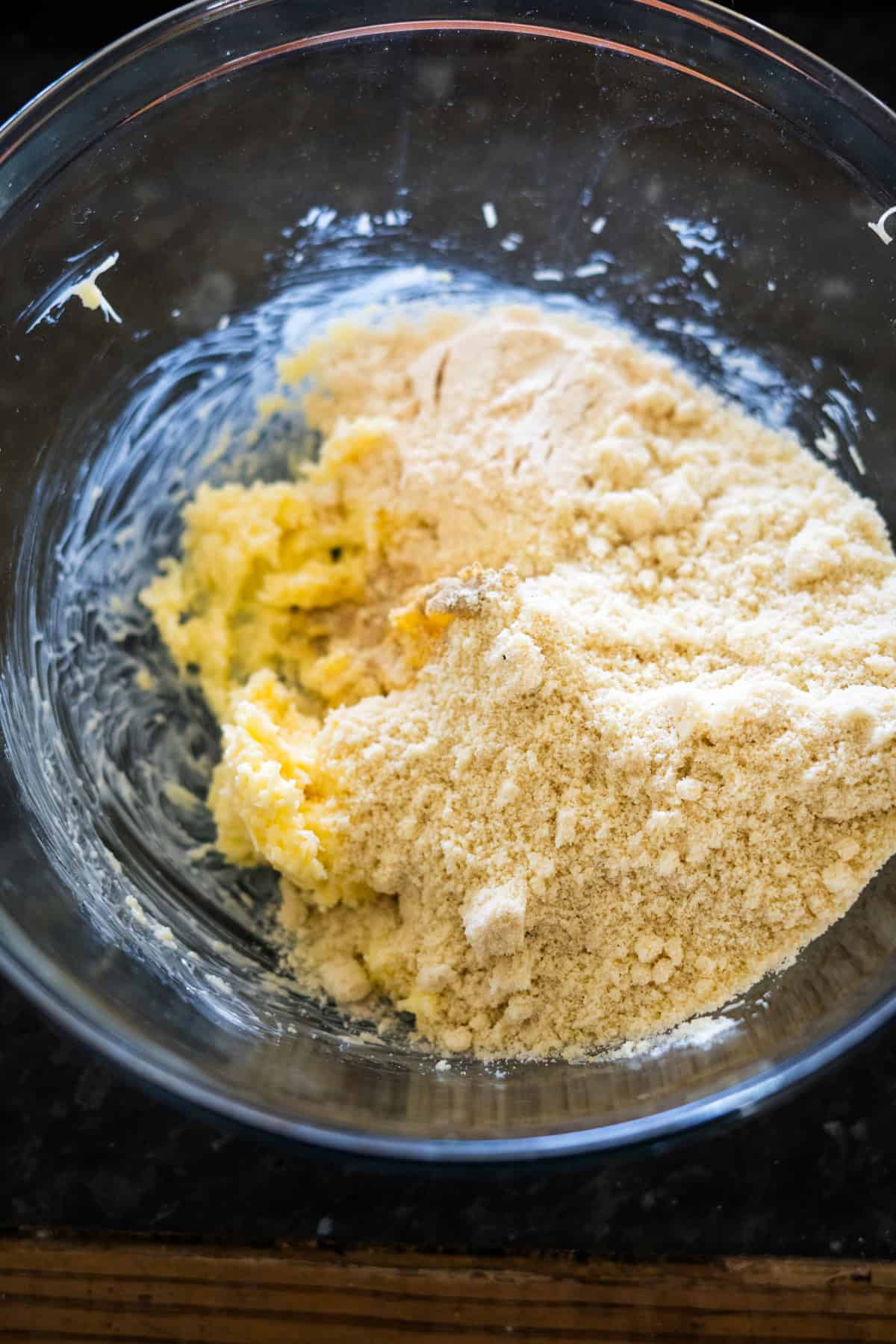 A glass bowl containing butter, almond flour, and the perfect hint of cheese crackers sits unmixed on a dark countertop.