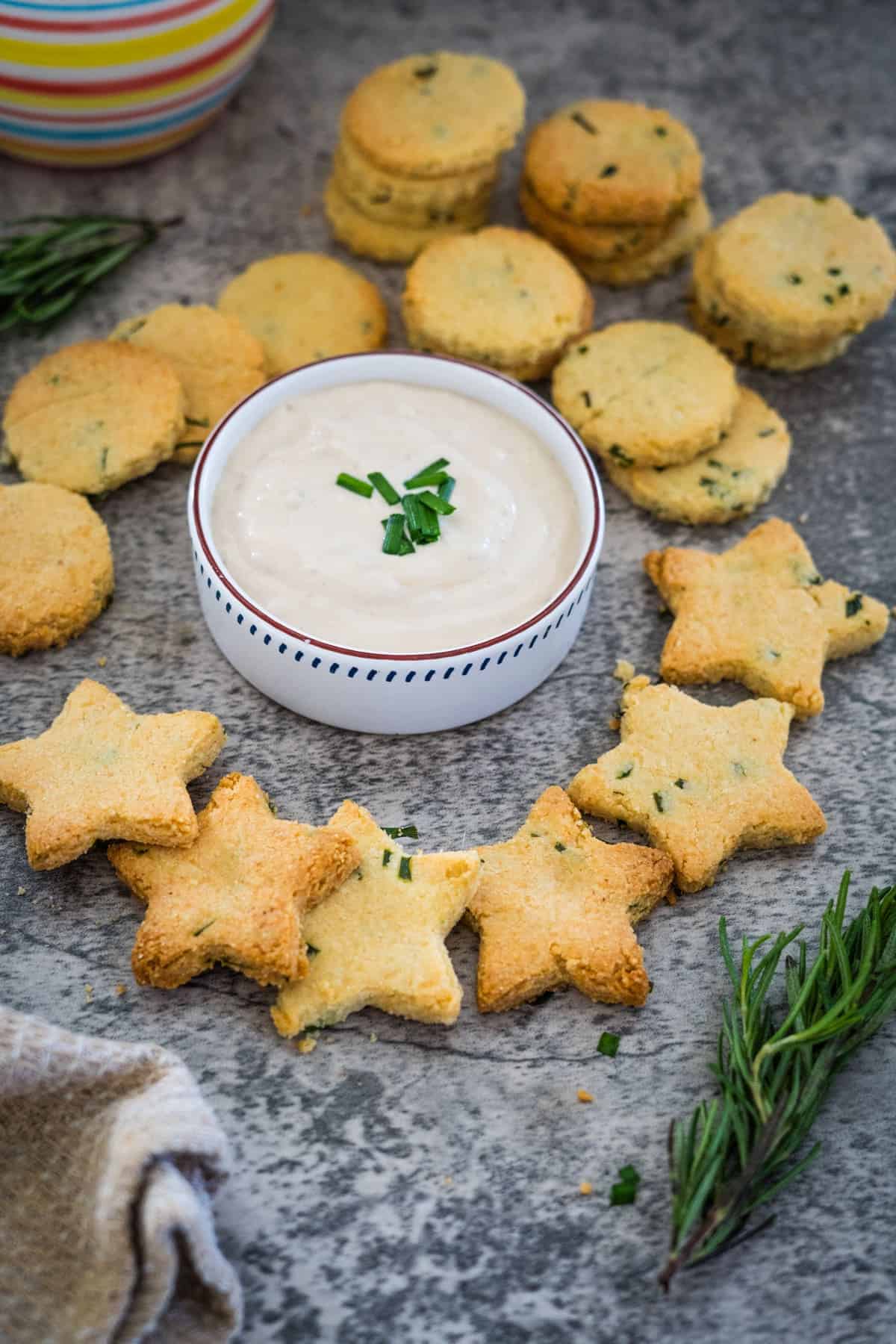 Star-shaped cheese crackers and round biscuits surround a bowl of creamy dip, garnished with herbs, on a stone surface. Rosemary sprigs and a cloth napkin are nearby.