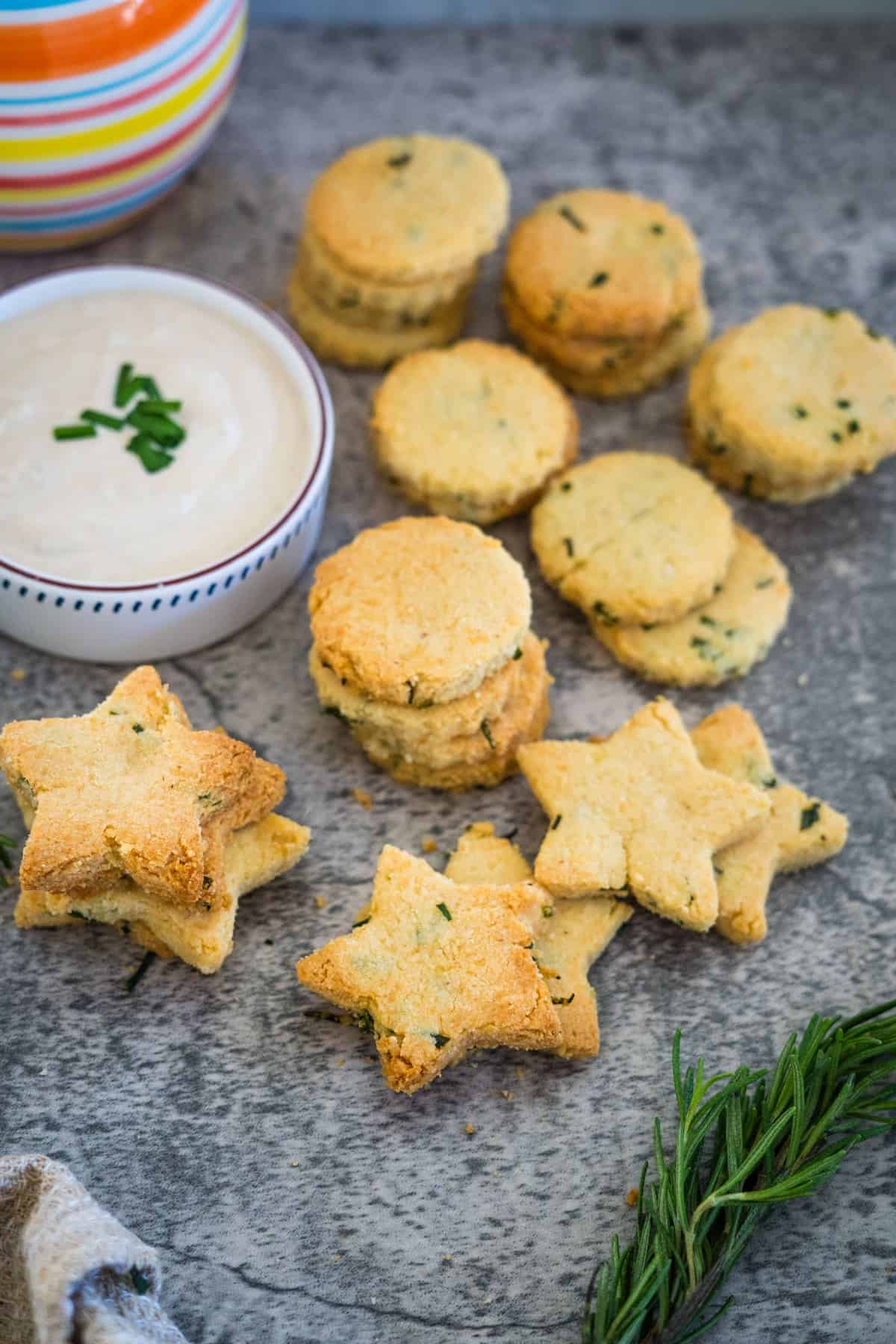 A variety of star and round-shaped herb and cheese crackers are displayed on a gray surface next to a bowl of creamy dip garnished with chives.