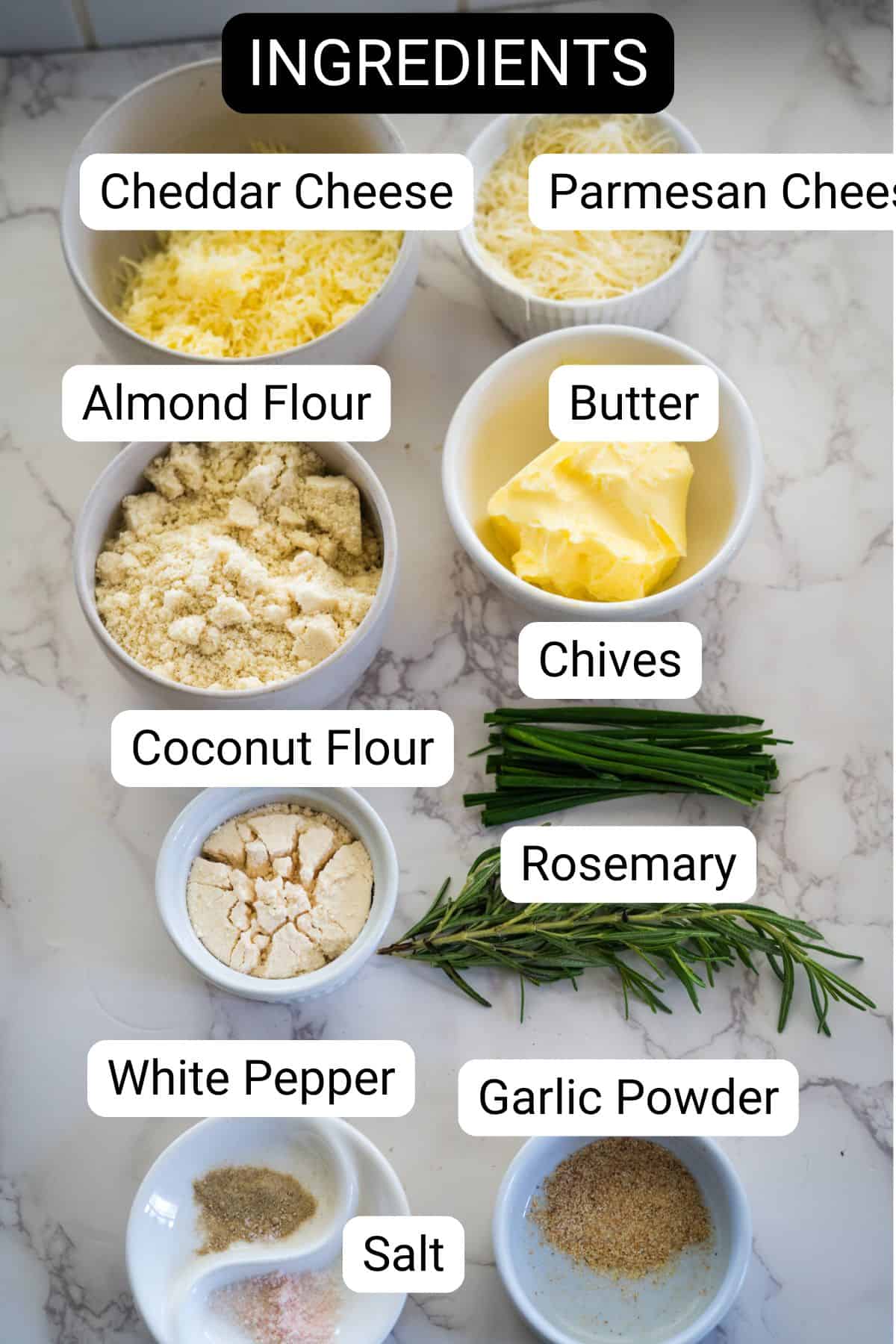 Ingredients on a table: cheddar cheese, parmesan cheese, almond flour, butter, chives, coconut flour, rosemary, white pepper, garlic powder, and salt. Perfect for crafting delicious homemade cheese crackers that offer a savory crunch with every bite.