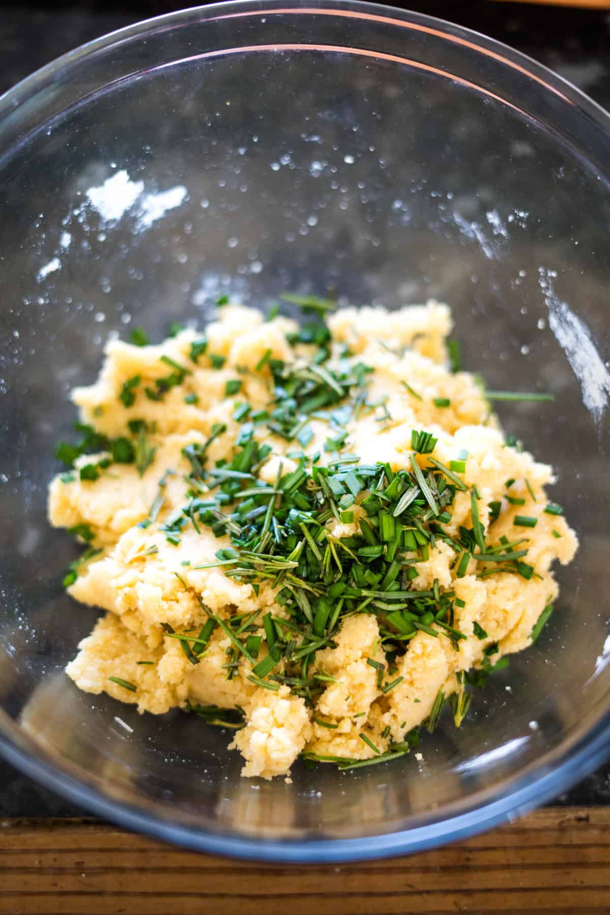 A glass bowl containing mashed potatoes topped with chopped fresh herbs and a sprinkle of crushed cheese crackers.