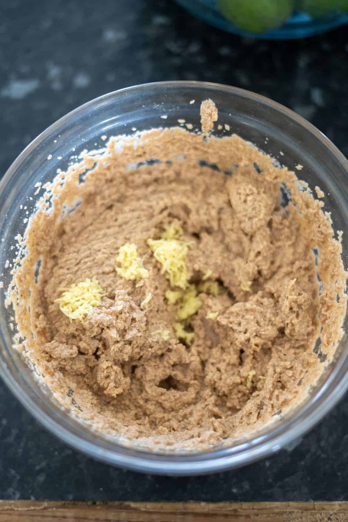 A glass bowl containing a mixture of brown ginger cake dough with grated ginger on top, set on a dark countertop.