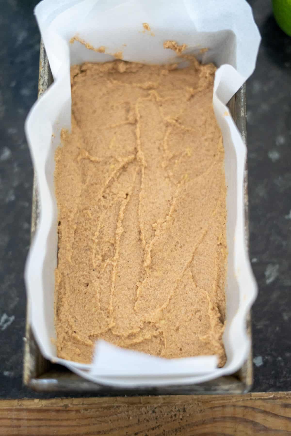 A loaf pan lined with parchment paper overflows with beige ginger cake batter, ready for baking on a dark countertop.