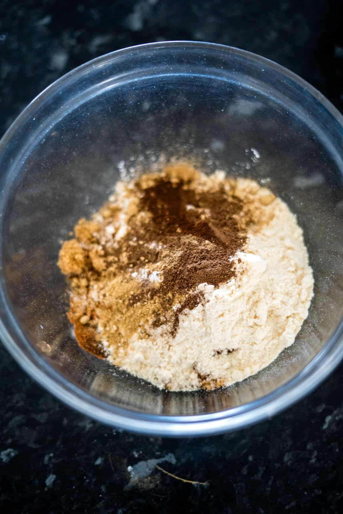 A mix of dry ingredients for a delicious ginger cake, including flour and spices, sits elegantly in a transparent glass bowl on a dark countertop.