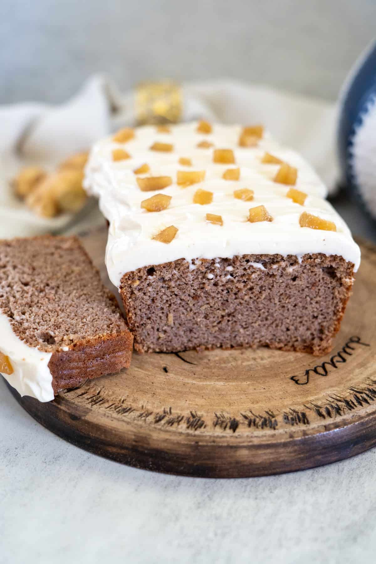 A sliced loaf of frosted ginger cake with small golden pieces on top is displayed on a round wooden board.