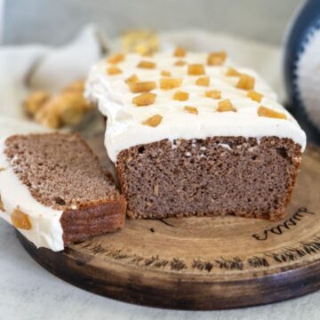 A sliced loaf of ginger cake is topped with white frosting and garnished with ginger pieces, all elegantly displayed on a wooden board.