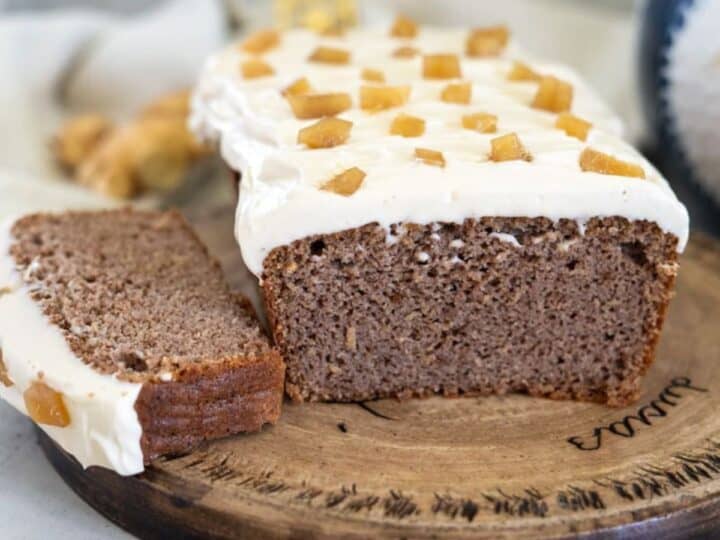 A sliced loaf of ginger cake is topped with white frosting and garnished with ginger pieces, all elegantly displayed on a wooden board.