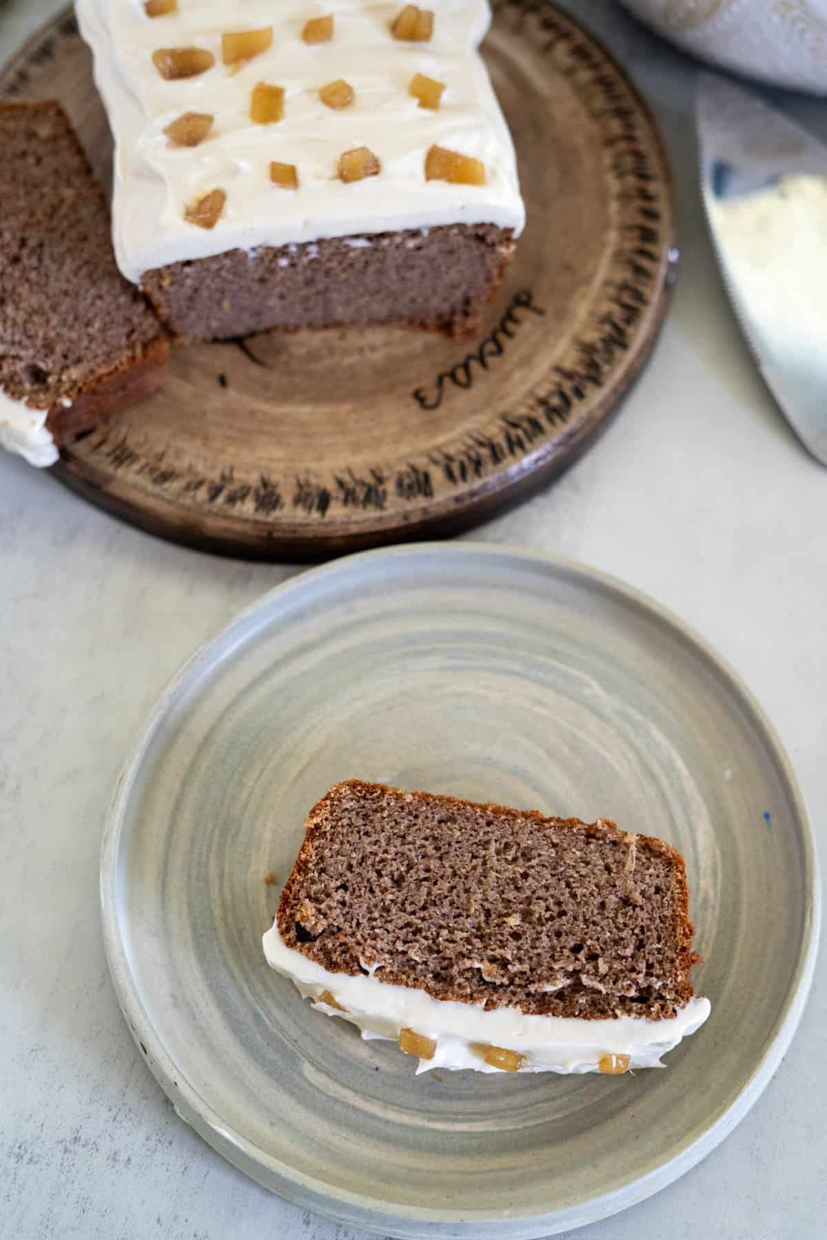 A slice of ginger cake with frosted icing sits on a plate, adorned with candied ginger pieces. In the background, the rest of the delicious cake rests on a wooden platter.