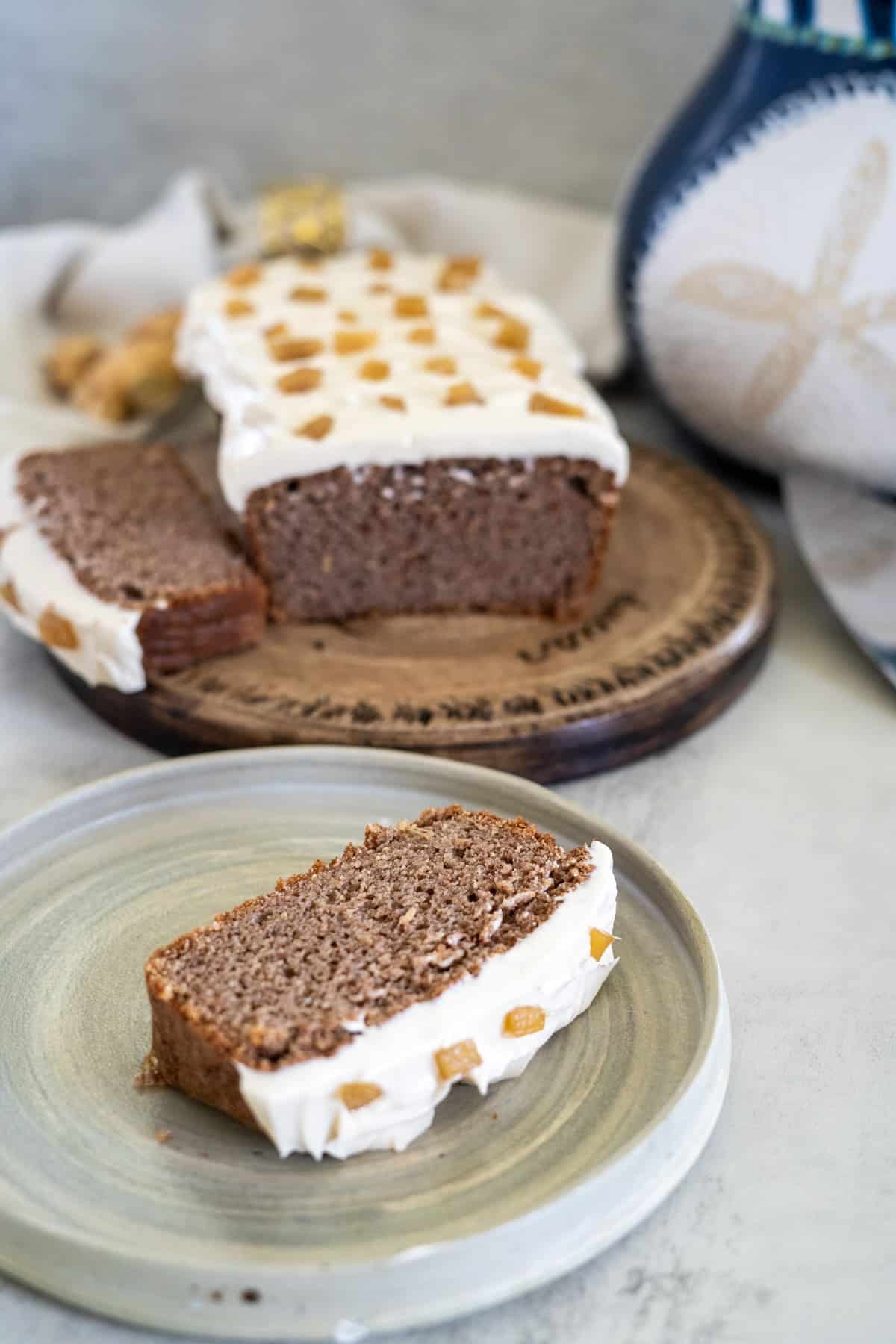 A slice of frosted ginger cake with candied ginger on top sits on a plate, in front of the rest of the loaf resting on a wooden board.