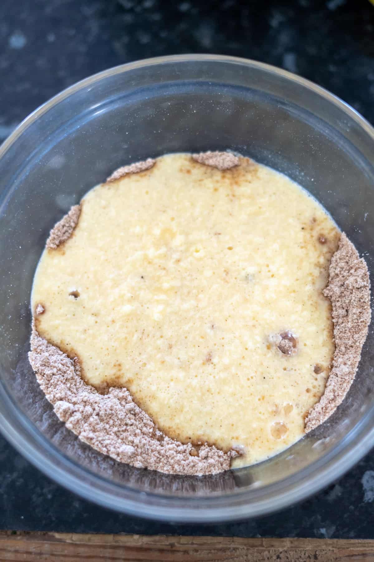 A glass bowl with a mixture of wet and dry ingredients partially combined for a ginger cake, sitting on a dark countertop.