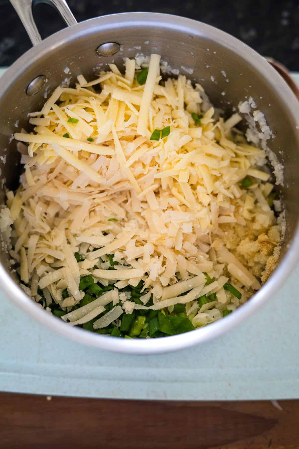 A metal pot containing shredded cheese, chopped green onions, and minced garlic, ideal ingredients for making delicious cauliflower croquettes.