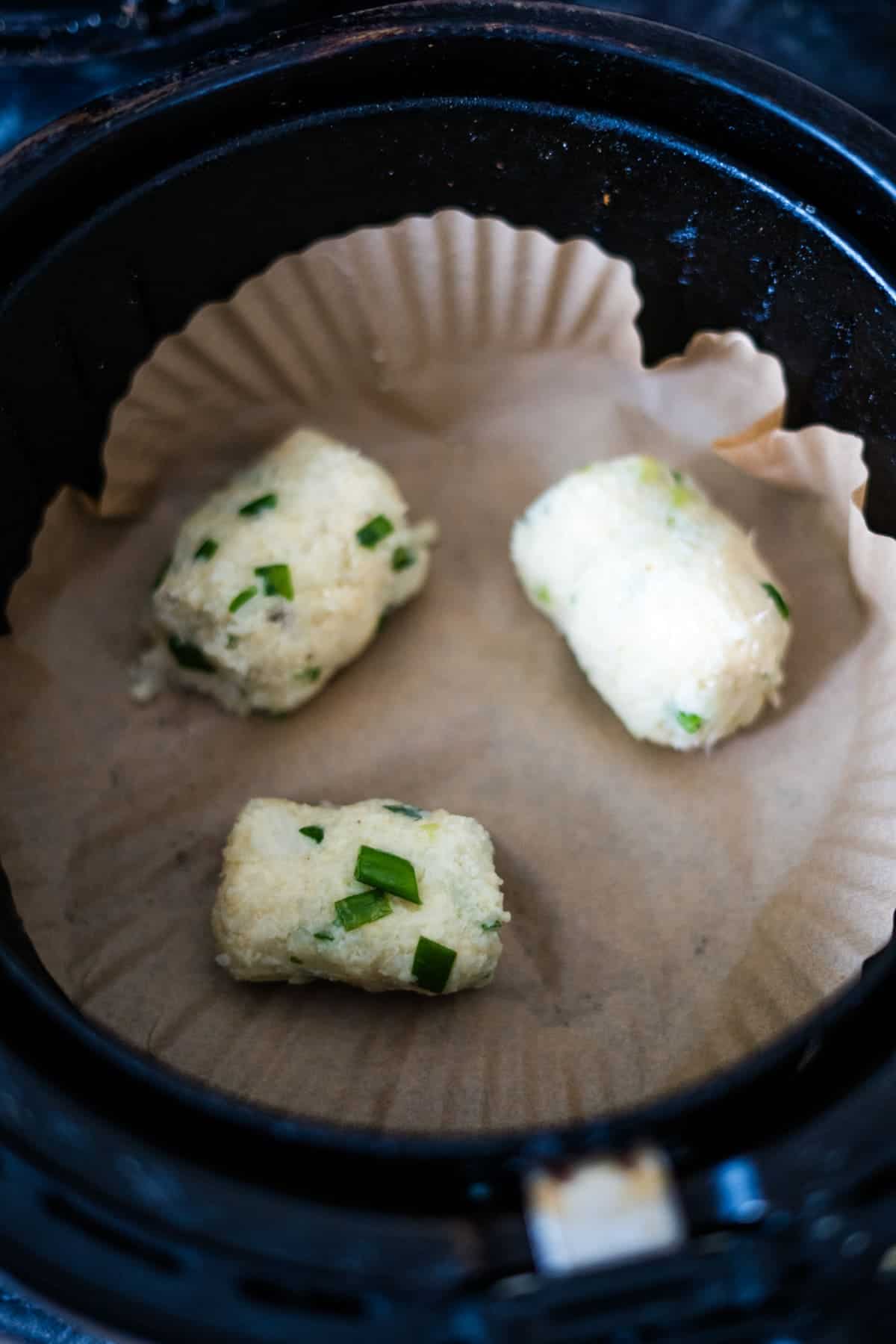 Three uncooked cauliflower croquettes with green herbs rest on parchment paper inside an air fryer basket.