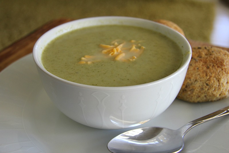 A bowl of green broccoli soup with shredded cheese on top, accompanied by a bread roll and a spoon on a white plate.