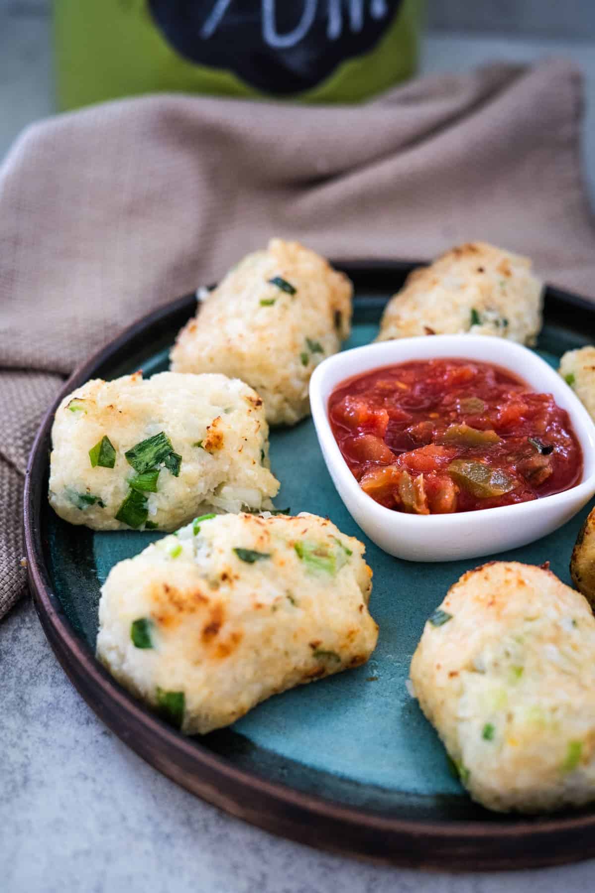 A plate of cauliflower croquettes arranged around a small bowl of tomato salsa, placed on a textured surface with a beige napkin in the background.