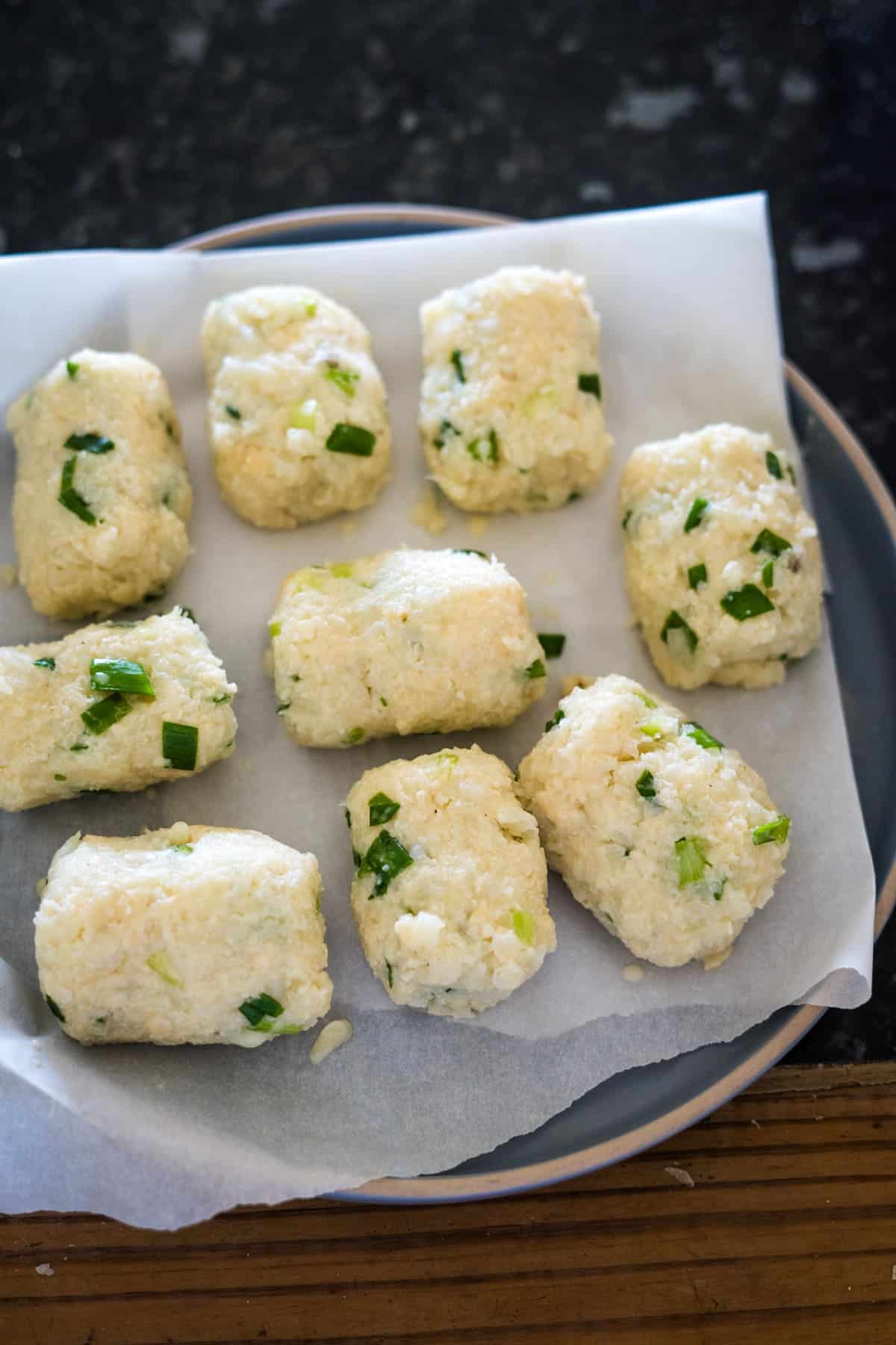 A plate of eight cauliflower croquettes garnished with chopped green onions sits on a piece of parchment paper.