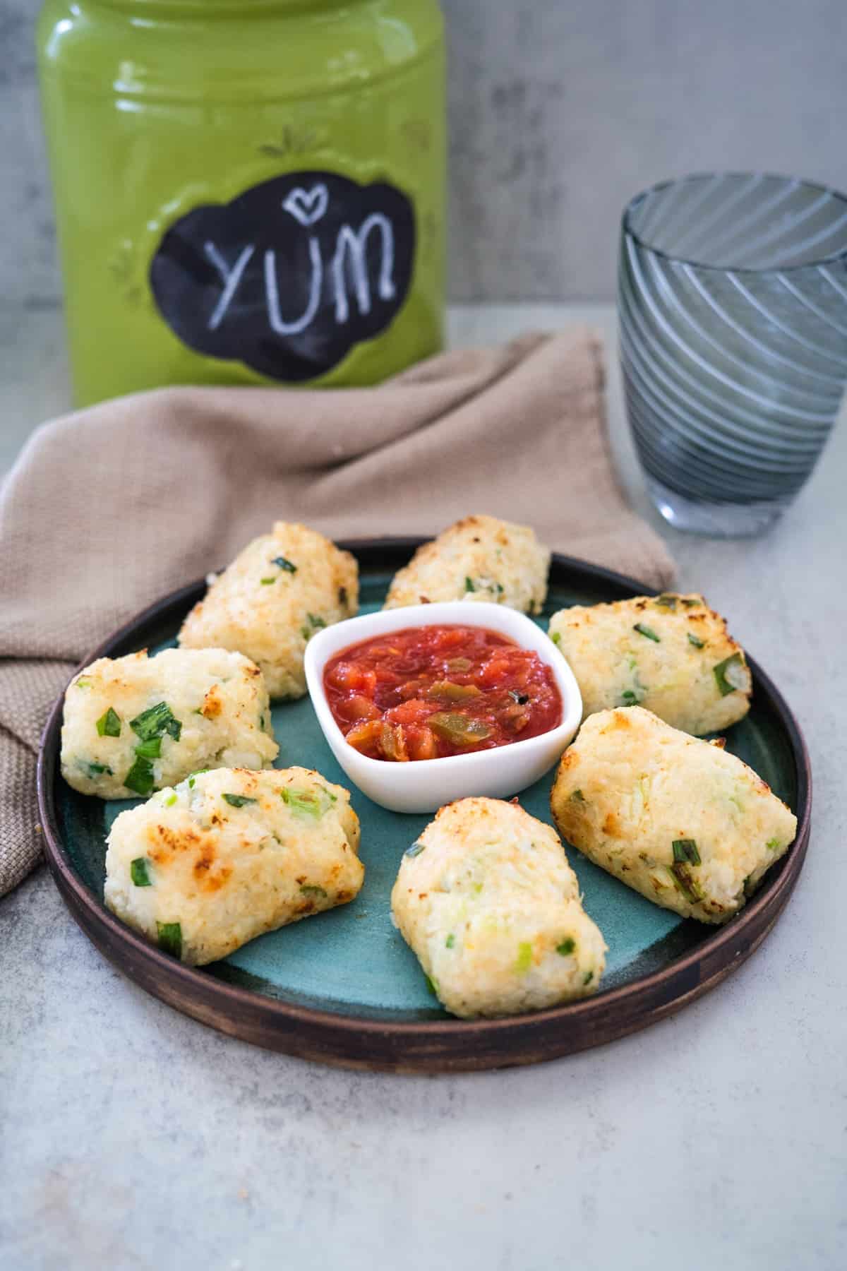 A plate of eight rectangular cauliflower croquettes with green flecks surrounds a small dish of red dip, placed on a table with a glass, napkin, and a green container in the background.