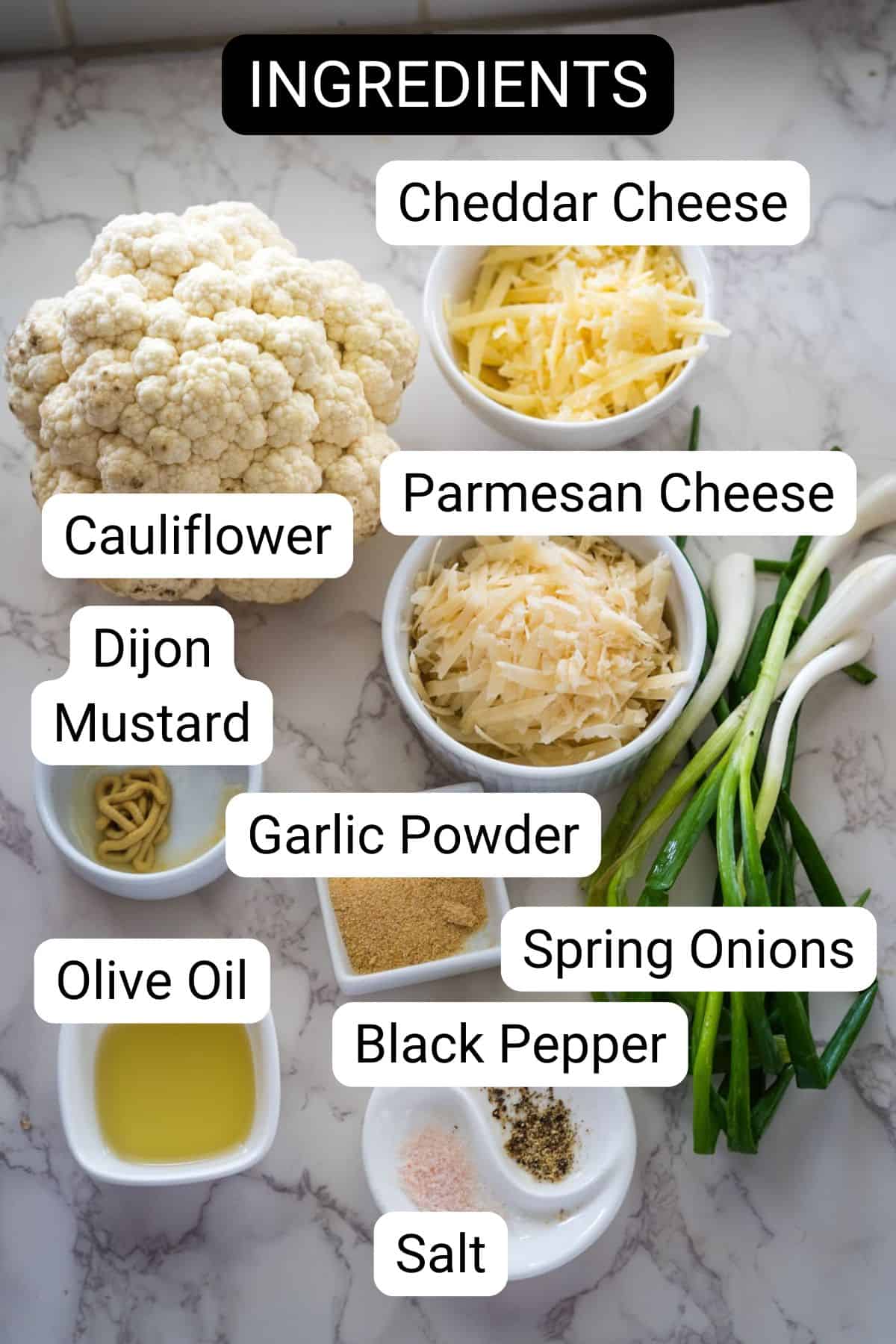 Ingredients for cauliflower croquettes on a marble counter: cauliflower, cheddar cheese, parmesan cheese, spring onions, dijon mustard, garlic powder, black pepper, salt, olive oil. Text labels identify each item.