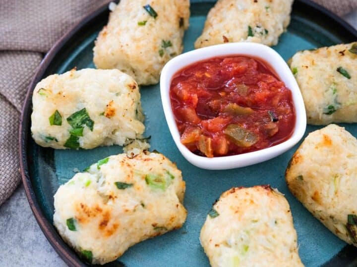 A plate with six golden-brown tater tots and cauliflower croquettes, garnished with green herbs, surrounding a small square dish of red salsa sauce. A beige cloth is in the background.