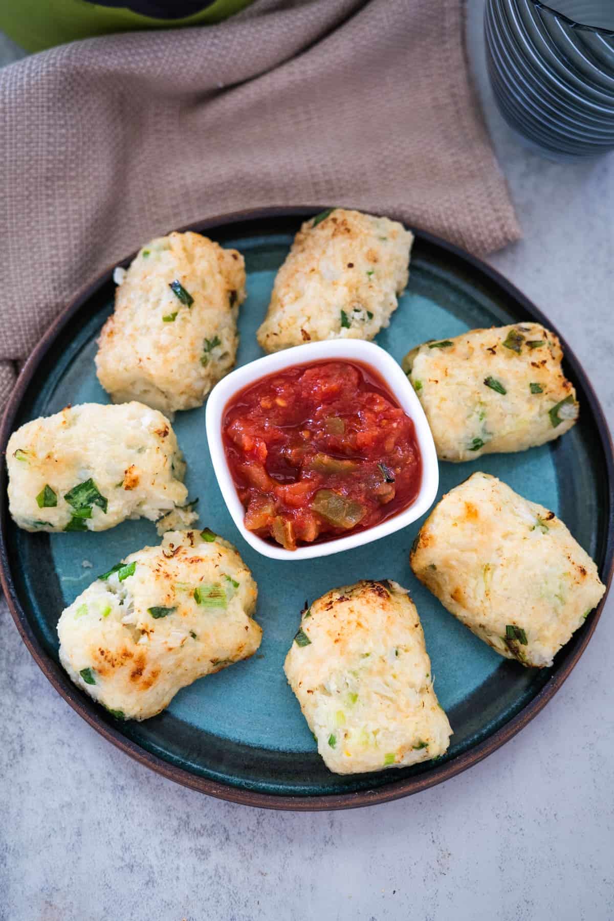 A blue plate with six rectangular-shaped cauliflower croquettes sprinkled with green herbs, surrounding a small square dish filled with red salsa.