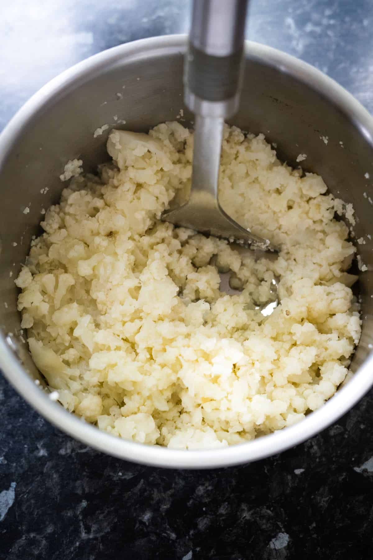 A pot filled with mashed cauliflower, alongside a metal masher resting in it, sits on a dark, speckled countertop—perfect for creating delicious cauliflower croquettes.