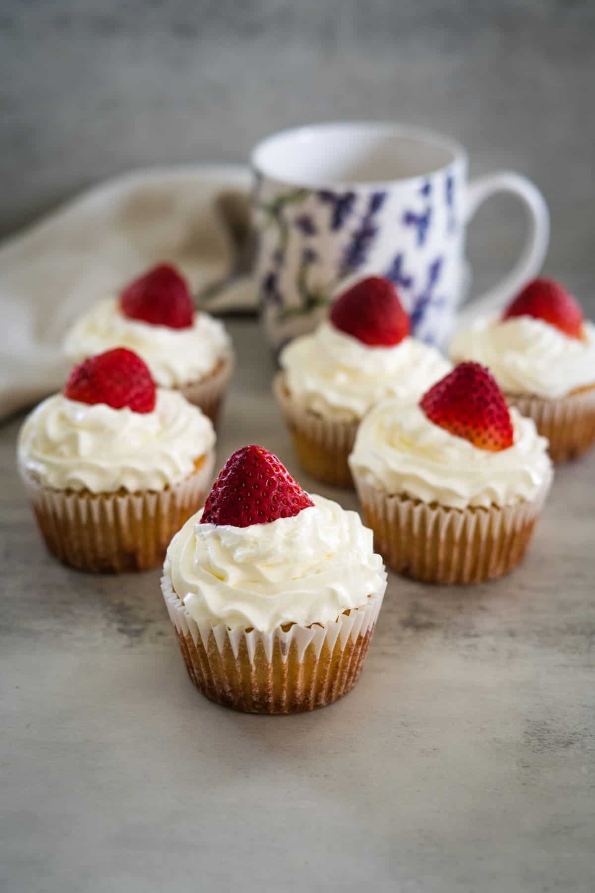 Six strawberry cream cupcakes topped with white frosting and a strawberry, arranged on a grey surface with a floral mug in the background.