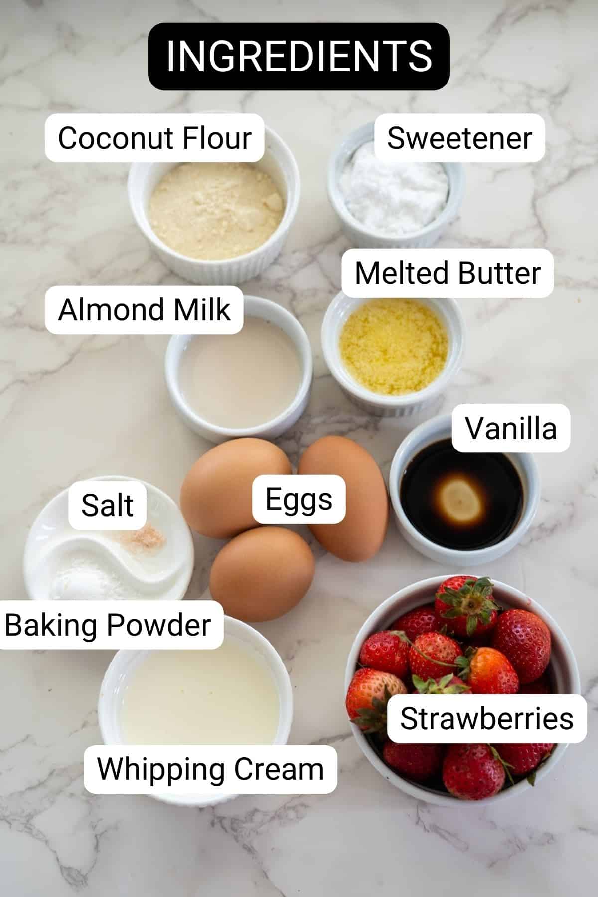 Ingredients for strawberry cream cupcakes laid out on a marble surface, including coconut flour, sweetener, almond milk, melted butter, vanilla, salt, baking powder, whipping cream, eggs, and strawberries.