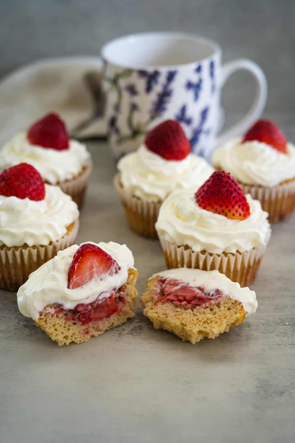 Several strawberry cream cupcakes topped with whipped cream and strawberries, one of which is cut in half to show the strawberry filling. A floral cup is in the background.