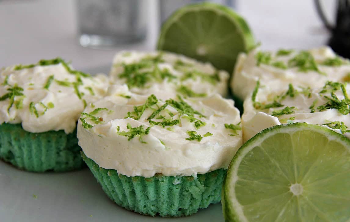 Key lime cupcakes with white frosting and lime zest, displayed with fresh lime slices, sit charmingly beside strawberry cream cupcakes.