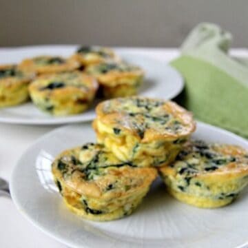 Small, round spinach and feta frittatas are arranged on two white plates, one in the foreground and one in the background. A green napkin is partially visible next to the plates.