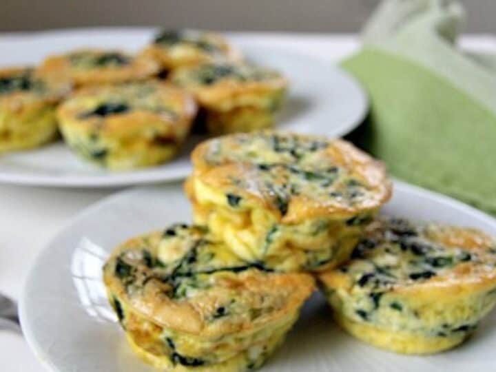 Small, round spinach and feta frittatas are arranged on two white plates, one in the foreground and one in the background. A green napkin is partially visible next to the plates.