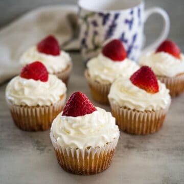 Six strawberry cream cupcakes topped with swirls of white frosting and whole strawberries. A floral patterned mug and a beige cloth are in the background.