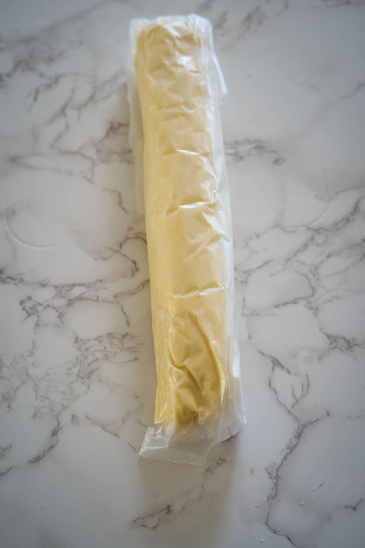A raw keto filo pastry roll wrapped in plastic, placed on a marble countertop.