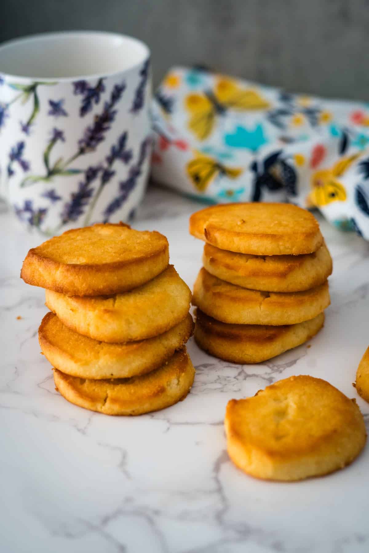 Stack of golden brown keto cream cheese cookies on a marble surface, with a floral mug and cloth in the background.