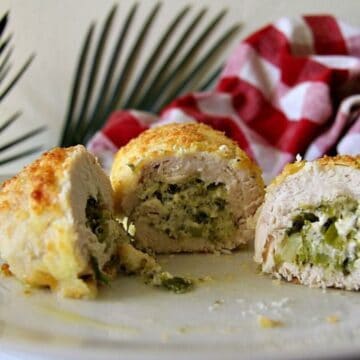 Close-up of succulent stuffed chicken breast sliced to reveal a delicious mixture of green vegetables and cheese, all presented on a white plate. A red and white checkered cloth and green plant leaves add a charming touch to the background.