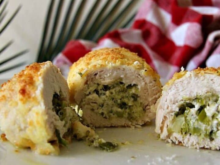 Close-up of succulent stuffed chicken breast sliced to reveal a delicious mixture of green vegetables and cheese, all presented on a white plate. A red and white checkered cloth and green plant leaves add a charming touch to the background.