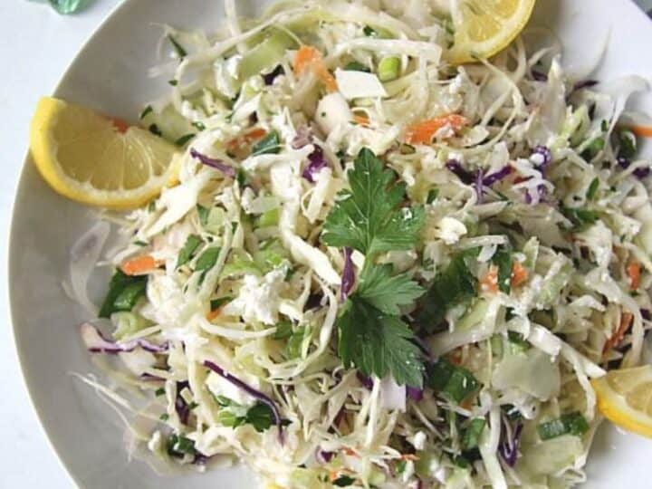 A white plate filled with a delightful coleslaw recipe made from shredded cabbage, carrots, and greens, garnished with lemon wedges and a parsley sprig.