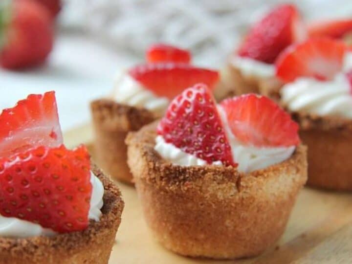 Mini cheesecakes topped with whipped cream and fresh strawberry slices, displayed on a wooden surface, inspired by keto strawberry shortcake.