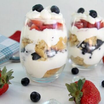 Two glass cups filled with layers of whipped cream, blueberries, strawberries, and cake pieces resemble a delightful cheesecake trifle. Fresh strawberries and blueberries are scattered on the white surface around the cups.