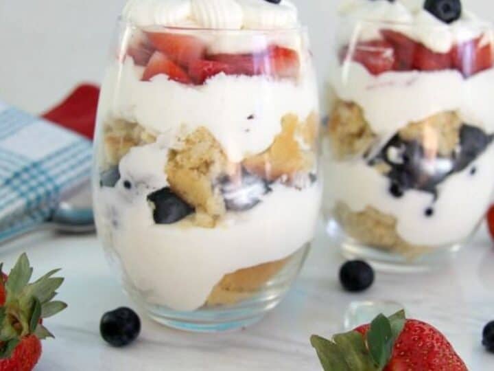 Two glass cups filled with layers of whipped cream, blueberries, strawberries, and cake pieces resemble a delightful cheesecake trifle. Fresh strawberries and blueberries are scattered on the white surface around the cups.