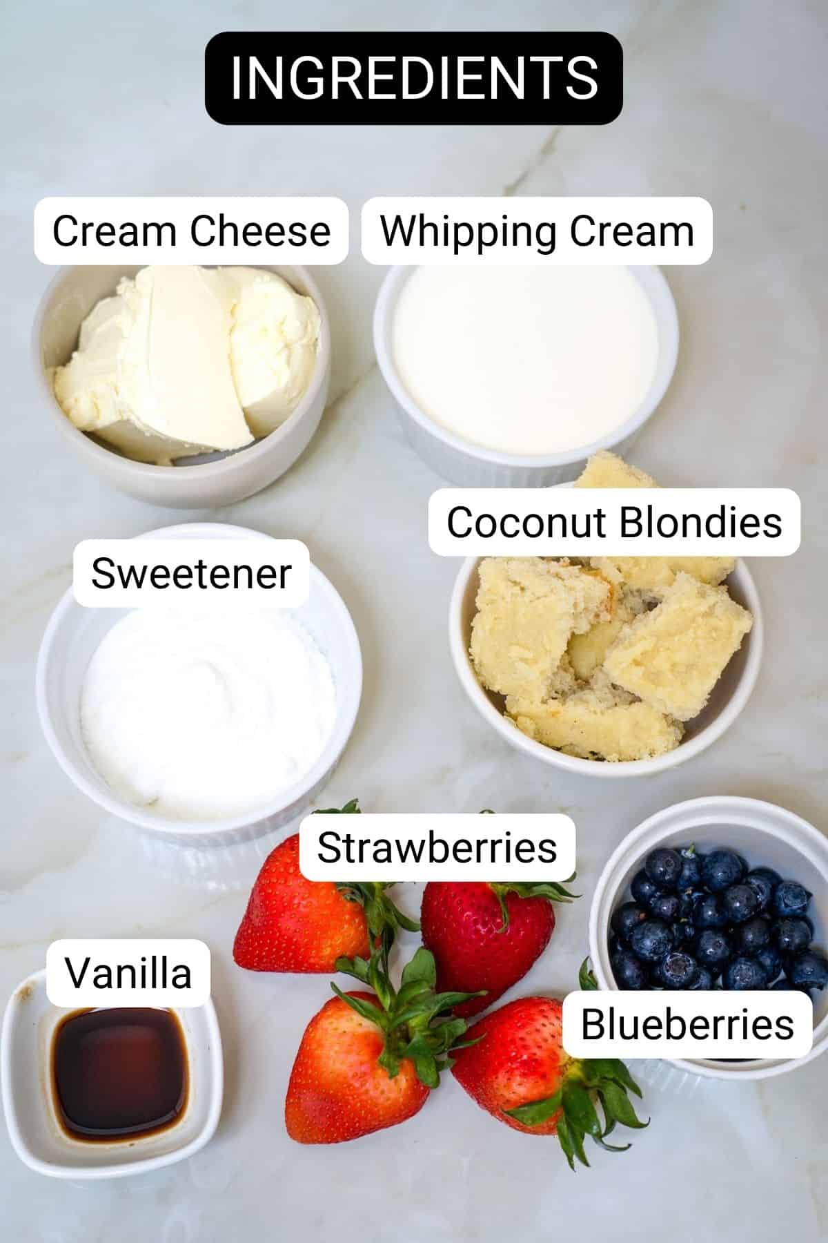 Ingredients on a counter: cream cheese, whipping cream, sweetener, coconut blondies, vanilla, strawberries, and blueberries—perfect for assembling a delicious cheesecake trifle. Each item is labeled with text.