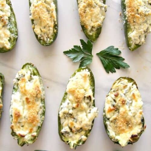Baked stuffed jalapeños filled with feta, topped with breadcrumbs, arranged on a white surface, accompanied by a parsley leaf.