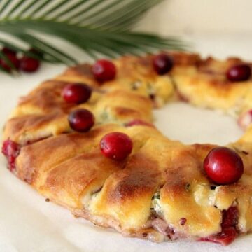 A wreath-shaped pastry filled with creamy cranberry brie and cranberries, topped with whole cranberries, rests on parchment paper. A palm leaf is visible in the background.