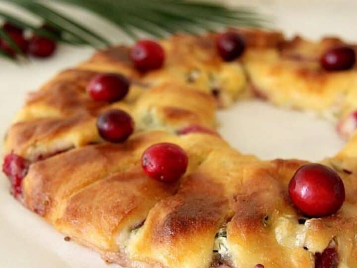 A wreath-shaped pastry filled with creamy cranberry brie and cranberries, topped with whole cranberries, rests on parchment paper. A palm leaf is visible in the background.