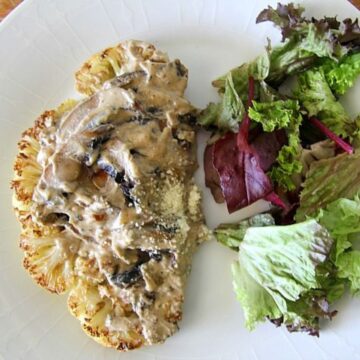 A plate with a grilled cauliflower steak topped with creamy mushroom sauce, served alongside a small mixed green salad featuring fresh ingredients.