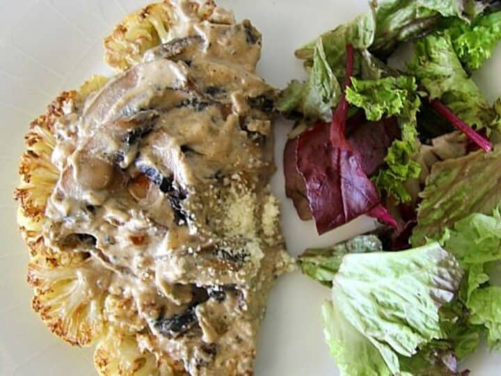 A plate with a grilled cauliflower steak topped with creamy mushroom sauce, served alongside a small mixed green salad featuring fresh ingredients.