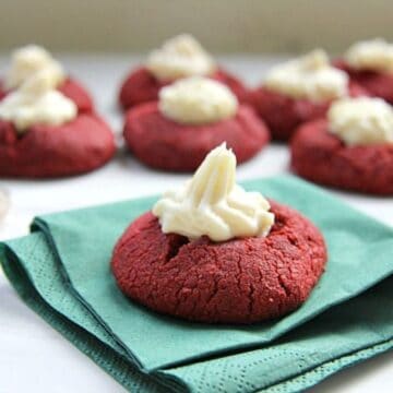 Red velvet cookies topped with cream cheese frosting are stacked on green napkins, with more of these delectable treats in the background.