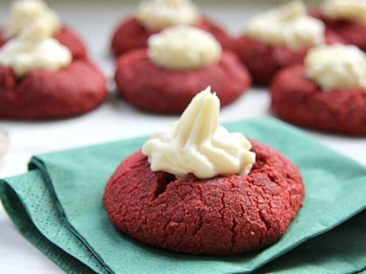 Red velvet cookies topped with cream cheese frosting are stacked on green napkins, with more of these delectable treats in the background.