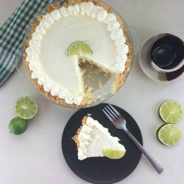 A keto key lime cheesecake with a slice cut out, topped with whipped cream and a lime wedge, next to whole limes, a checkered cloth, and a cup of coffee on a white surface. A slice is served on a black plate.