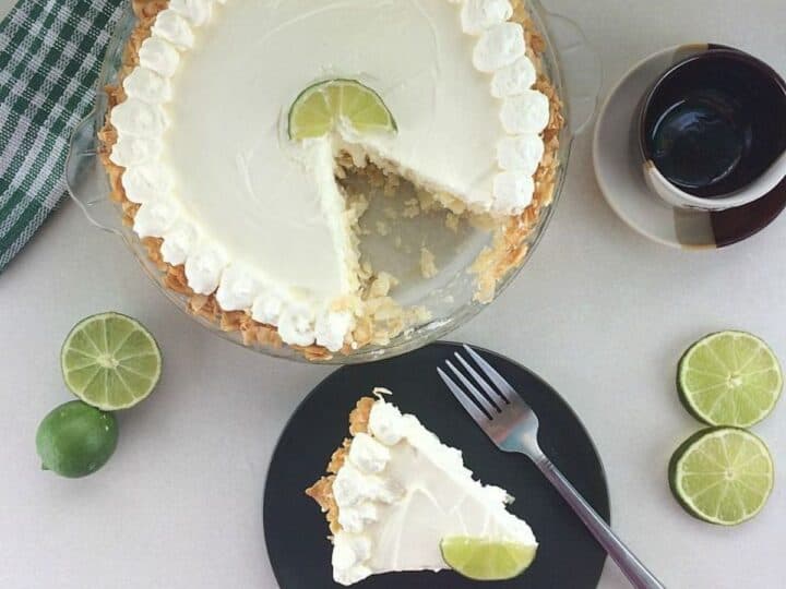 A keto key lime cheesecake with a slice cut out, topped with whipped cream and a lime wedge, next to whole limes, a checkered cloth, and a cup of coffee on a white surface. A slice is served on a black plate.